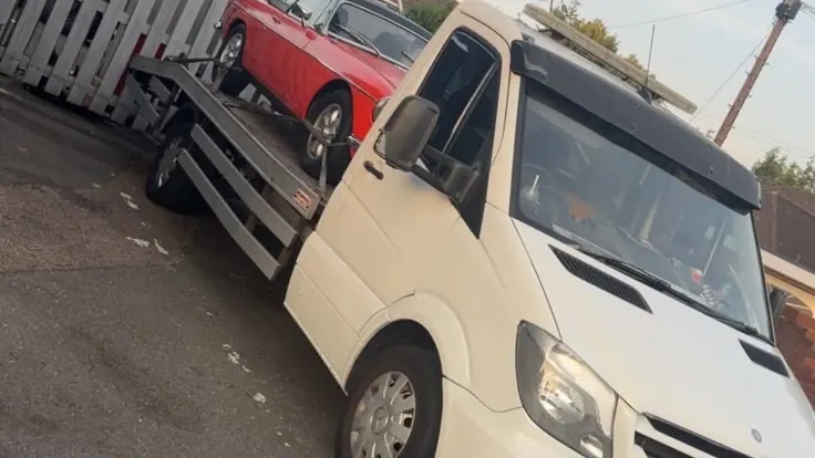 White recovery truck carrying a red classic car, parked beside a wooden fence