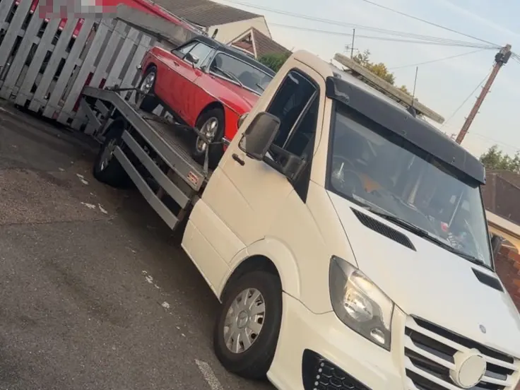 White recovery truck carrying a red classic car, parked beside a wooden fence