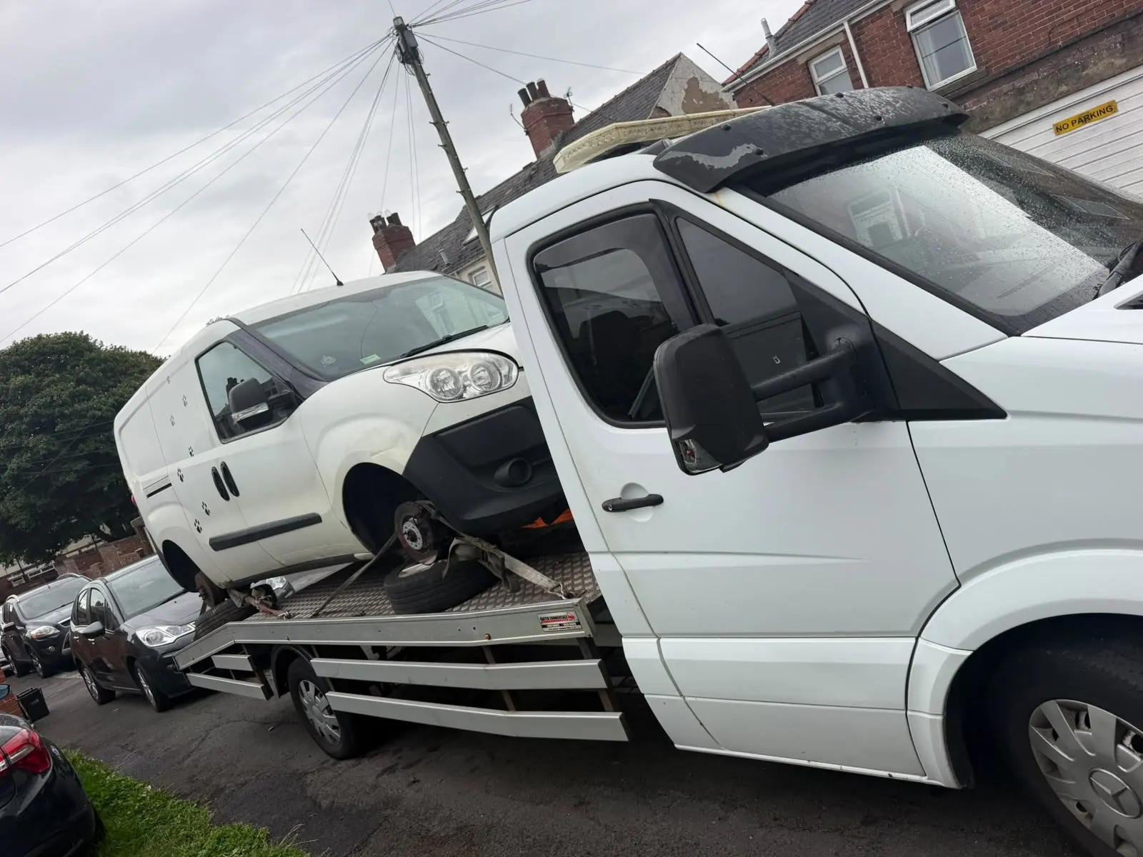 White recovery truck carrying a small white van with a front wheel off, on a street of red-brick terraced houses