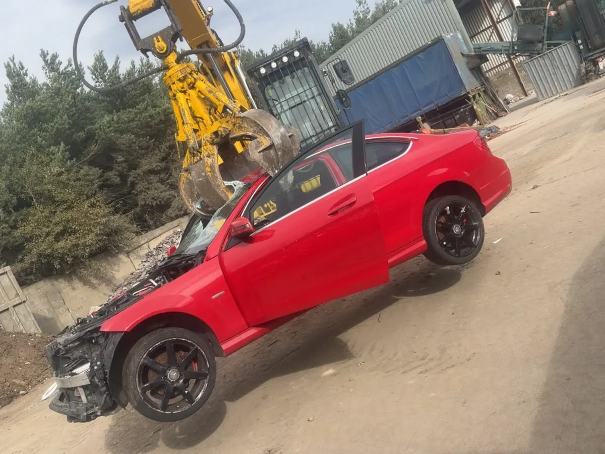 Damaged red coupé on the ground at a scrapyard, next to a yellow grab