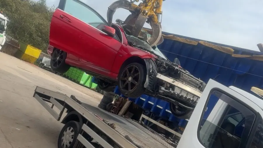 Yellow grab at a scrapyard lifting a damaged red car off the back of the recovery truck