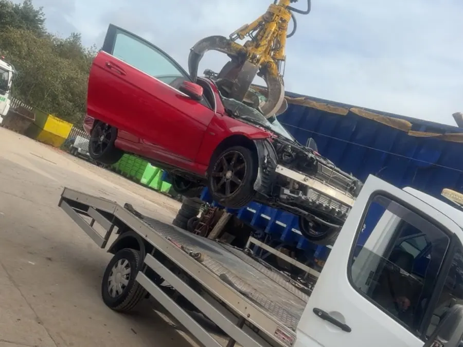 Yellow grab at a scrapyard lifting a damaged red car off the back of the recovery truck